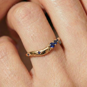 Puede incluir: Un anillo dorado curvado que presenta una serie de pequeñas piedras preciosas redondas de color azul intenso engastadas a lo largo de la banda. La superficie metálica pulida incluye detalles grabados en forma de estrella entre las piedras.