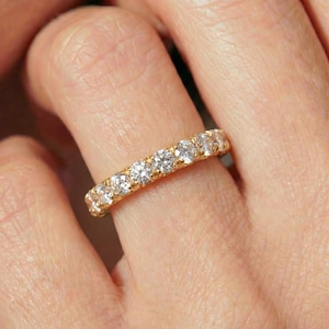 May include: A gold eternity band featuring a continuous row of round, brilliant-cut clear stones set in a shared-prong setting. The ring encircles the finger, showcasing a polished yellow gold metal band with multiple sparkling white gemstones.