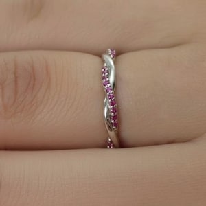 May include: A silver-colored ring with a twisted design, adorned with small, vibrant pink gemstones. The ring is worn on a finger, showcasing its intricate details and sparkling stones. The ring's design is a continuous, interwoven pattern.