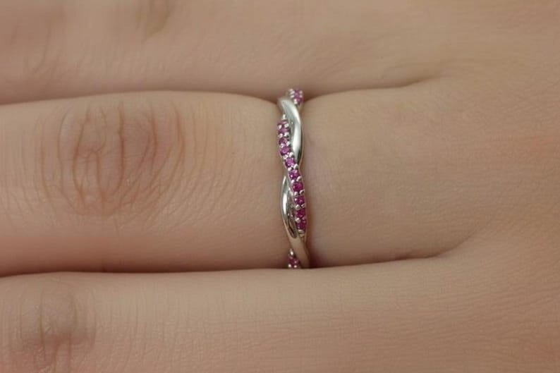 May include: A silver-colored ring with a twisted design, adorned with small, vibrant pink gemstones. The ring is worn on a finger, showcasing its intricate details and sparkling stones. The ring's design is a continuous, interwoven pattern.