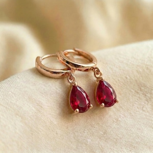 May include: Rose gold hoop earrings with teardrop-shaped, deep red gemstones. The earrings are displayed on a cream-colored fabric, showcasing the jewelry's elegant design and vibrant color. The gemstones have a faceted cut.