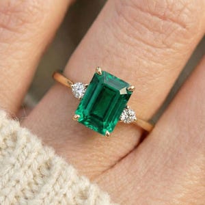 May include: A gold ring featuring a large emerald-cut green gemstone, flanked by two smaller, round, clear gemstones. The ring is set on a slender gold band. The ring is displayed on a person's finger, with a cream-colored sweater in the background.