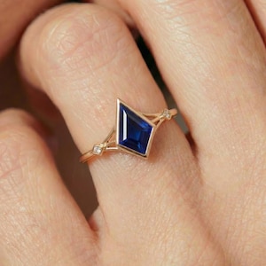 May include: A gold ring featuring a geometric, kite-shaped, deep blue gemstone. The ring has a delicate design with small accent stones on either side of the main stone. The ring is worn on a finger.