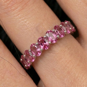 Puede incluir: Un anillo de plata con una serie de piedras preciosas rosas de corte ovalado. Las piedras están engastadas juntas, creando una banda continua de color. El anillo se muestra en un dedo, resaltando el diseño y el brillo de las piedras.