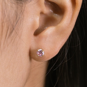 May include: A close-up of a person's ear with a small pink gemstone earring. The earring is set in gold and has a simple, classic design.