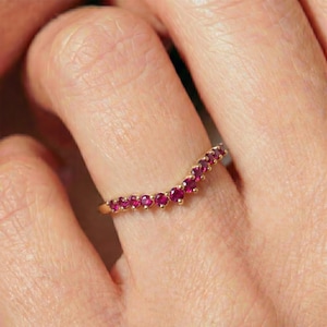 May include: A gold ring with a curved design, set with small, vibrant red gemstones. The ring is shown on a finger, highlighting the delicate setting and the rich color of the stones. The ring is a piece of jewelry.