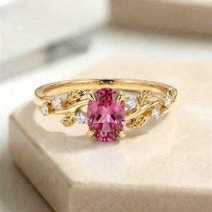 Puede incluir: Un anillo dorado con una piedra preciosa rosa ovalada como pieza central. El anillo está adornado con diseños en forma de hojas y pequeñas piedras preciosas transparentes. La banda tiene una estética delicada inspirada en la naturaleza.