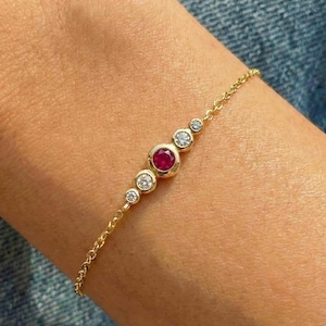 Puede incluir: Una delicada pulsera de oro con una serie de piedras preciosas. La pulsera tiene un rubí rojo redondo central, flanqueado por cuatro diamantes redondos y transparentes más pequeños. La pulsera está en el brazo de una persona.