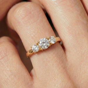 May include: A gold ring with three clear, round-cut gemstones. The center stone is larger than the two flanking stones. The ring is set on a finger, showcasing the design and sparkle of the gemstones.