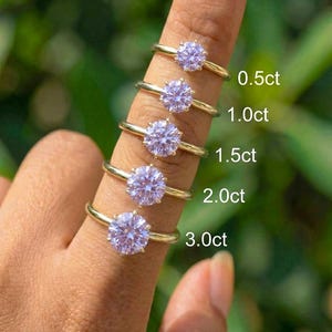 May include: Five gold rings with round, light purple gemstones are displayed on a finger. The rings are labeled with carat weights: 0.5ct, 1.0ct, 1.5ct, 2.0ct, and 3.0ct. The background is a blurred green.
