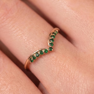 May include: A V-shaped ring crafted from rose gold, featuring a series of small, round, vibrant green gemstones set along the top curve of the band. The design follows a chevron silhouette, highlighting the polished metal and the stones.