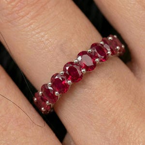 May include: A silver ring featuring a series of oval-cut, vibrant red gemstones. The gemstones are set closely together, creating a continuous band of color. The ring is shown on a person's finger, highlighting the jewelry's design and sparkle.