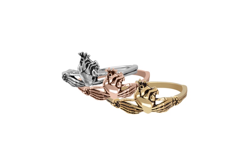 Puede incluir: Tres anillos de Claddagh en plata, oro rosa y oro. Cada anillo presenta un coraz&oacute;n sostenido por dos manos, coronado por una corona. Los anillos se muestran sobre un fondo blanco.