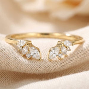 Op de afbeelding: Een goudkleurige ring met een uniek ontwerp. De ring heeft een reeks marquise-geslepen en ronde heldere edelstenen die aan weerszijden van de band zijn gegroepeerd, waardoor een bloem- of bladachtig uiterlijk ontstaat. De ring is geplaatst tegen een neutrale stoffen achtergrond.