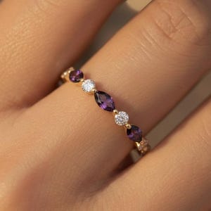 Op de afbeelding: Een gouden ring met afwisselend marquise-geslepen paarse edelstenen en ronde witte edelstenen. De ring heeft kleine accentstenen en wordt op een vinger getoond.