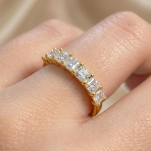 Könnte beinhalten: Ein goldener Ring mit einer Reihe quadratisch geschliffener, klarer Edelsteine. Der Ring hat ein gelbgoldenes Band, wobei die Edelsteine in einer Reihe angeordnet sind. Der Ring wird an einem Finger gezeigt.