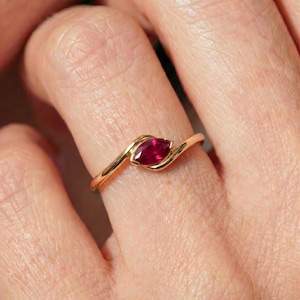May include: A gold ring featuring a single marquise-cut deep red gemstone set in a bypass style band. The metal band has a smooth, polished finish that curves around the central stone, creating a sleek and continuous look on the finger.