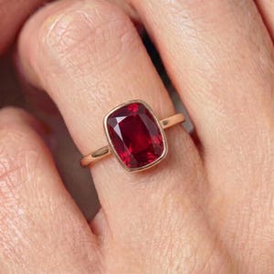 May include: A ring featuring a cushion-cut deep red gemstone set in a polished gold band. The stone is secured by a bezel setting that frames the faceted surface, highlighting the vibrant crimson colour and the metallic lustre of the gold band.
