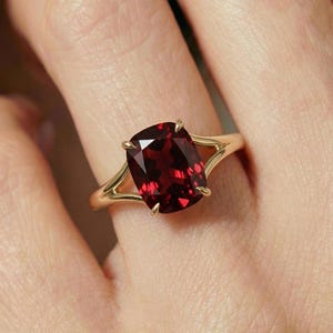 Puede incluir: Un anillo de oro con una gran gema roja oscura con talla cojín. La banda del anillo tiene un diseño dividido, y la gema está sujeta por cuatro garras. El anillo se muestra en un dedo.