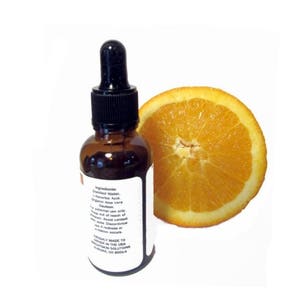 May include: A brown glass bottle with a black dropper and a white label, next to a sliced orange. The label lists ingredients and cautions. The orange slice is bright orange and shows the fruit's texture. The bottle appears to contain a liquid serum.