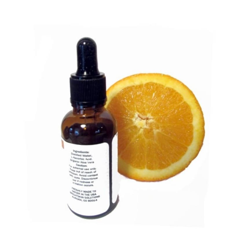 May include: A brown glass bottle with a black dropper and a white label, next to a sliced orange. The label lists ingredients and cautions. The orange slice is bright orange and shows the fruit's texture. The bottle appears to contain a liquid serum.