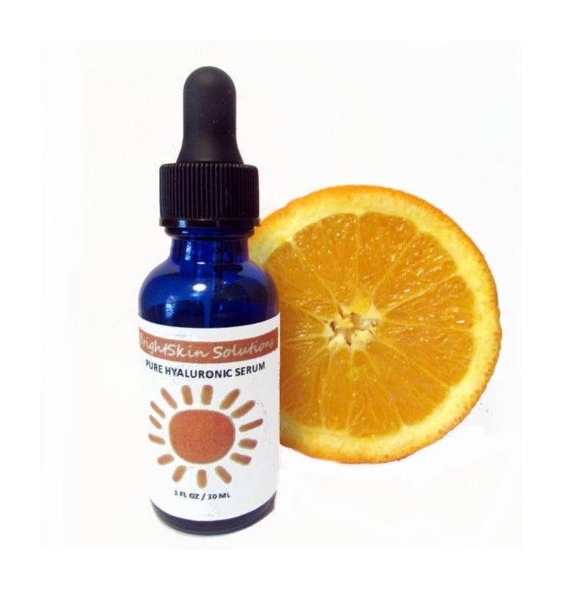 May include: A blue glass bottle of BrightSkin Solutions Pure Hyaluronic Serum with a black dropper and a white label featuring a sun design. A slice of orange is next to the bottle. The bottle contains 1 fl oz / 30 ml.