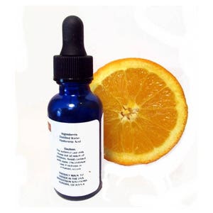 May include: A cobalt blue glass bottle with a black dropper, next to a sliced orange. The bottle has a white label with text that reads "Ingredients: Distilled Water, Hyaluronic Acid." The orange slice is bright orange.