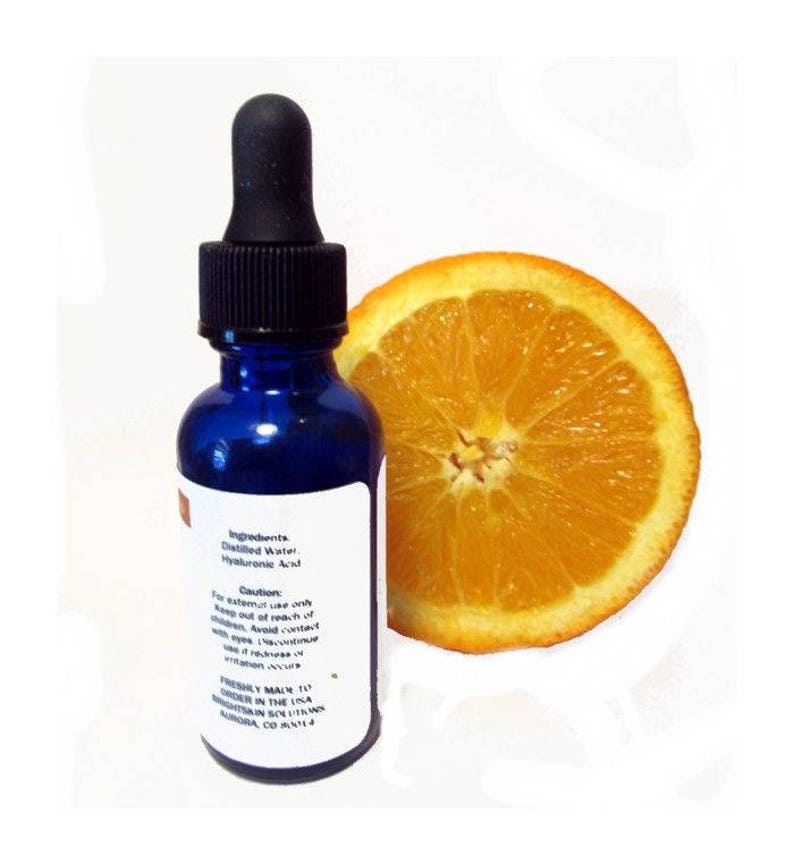 May include: A cobalt blue glass bottle with a black dropper, next to a sliced orange. The bottle has a white label with text that reads "Ingredients: Distilled Water, Hyaluronic Acid." The orange slice is bright orange.