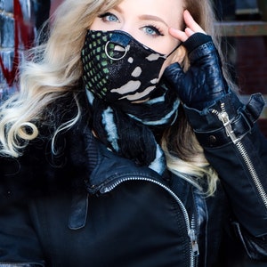 May include: A woman wearing a black leather jacket with a zip, a black and green patterned face mask with a silver ring, and a blue and black scarf.