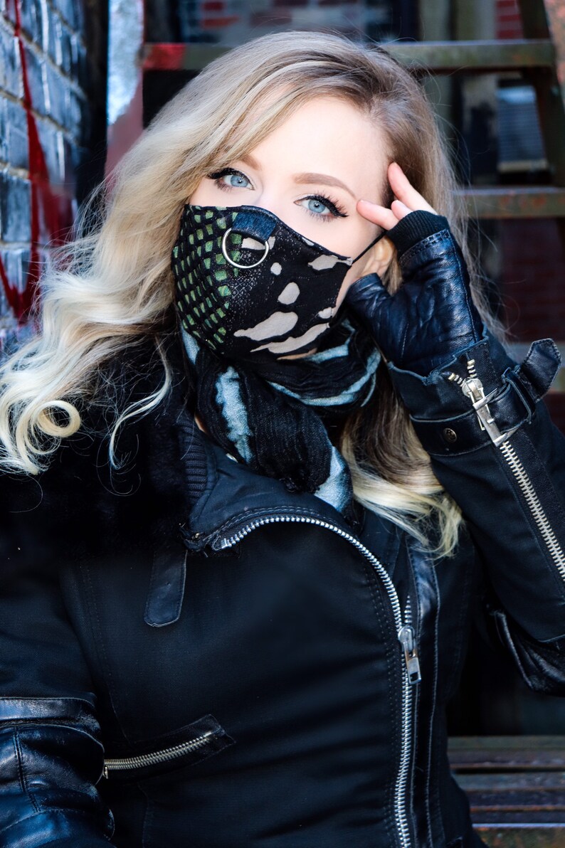 May include: A woman wearing a black leather jacket with a zip, a black and green patterned face mask with a silver ring, and a blue and black scarf.