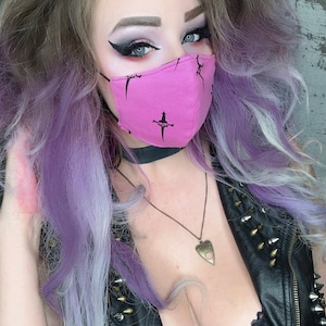 May include: A woman with purple and blonde hair is wearing a black choker, a black leather vest with silver studs, and a pink face mask with a black cross design. She is also wearing a necklace with a pendant in the shape of a  ouija board.
