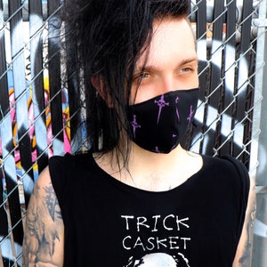 May include: A person with black hair wearing a black sleeveless shirt with the text "TRICK CASKET" and a skull graphic. They are wearing a black face mask with a purple skull and crossbones design.