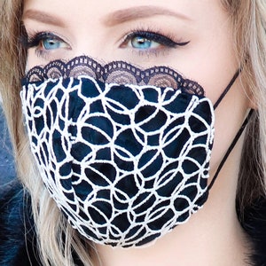 Może przedstawiać: Czarna maska ​​na twarz z białymi koronkami. Maska ​​ma geometryczny wz&oacute;r i jest wykonana z miękkiej, oddychającej tkaniny.