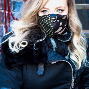 May include: A woman with long blonde hair wearing a black jacket with a fur collar and a black and green patterned face mask with a silver ring.