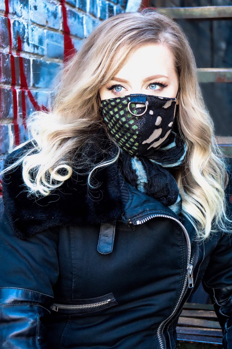 May include: A woman with long blonde hair wearing a black jacket with a fur collar and a black and green patterned face mask with a silver ring.