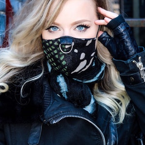 May include: A woman with blonde hair wearing a black leather jacket and a black and green patterned face mask with a silver ring.