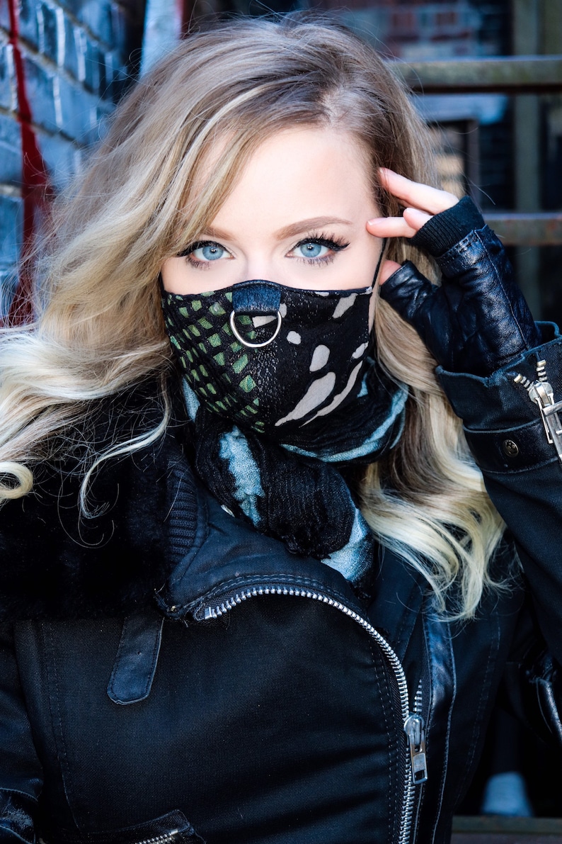 May include: A woman with blonde hair wearing a black leather jacket and a black and green patterned face mask with a silver ring.