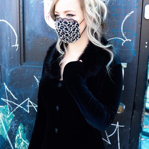 Może przedstawiać: Kobieta w czarnej aksamitnej sukience z kołnierzem z futra i czarno-białą maską na twarz. Stoi przed niebiesko-czarną drzwiami z białymi kredowymi oznaczeniami.