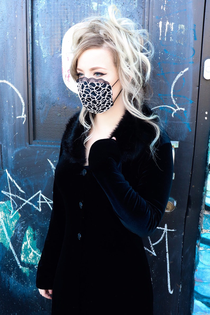 Może przedstawiać: Kobieta w czarnej aksamitnej sukience z kołnierzem z futra i czarno-białą maską na twarz. Stoi przed niebiesko-czarną drzwiami z białymi kredowymi oznaczeniami.