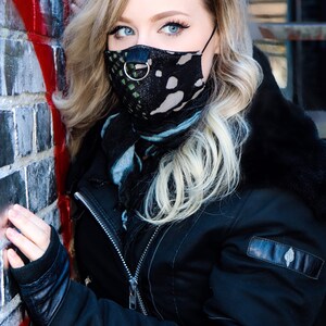 May include: A woman wearing a black leather jacket with a black and green patterned face mask. The mask has a silver ring attached to it.