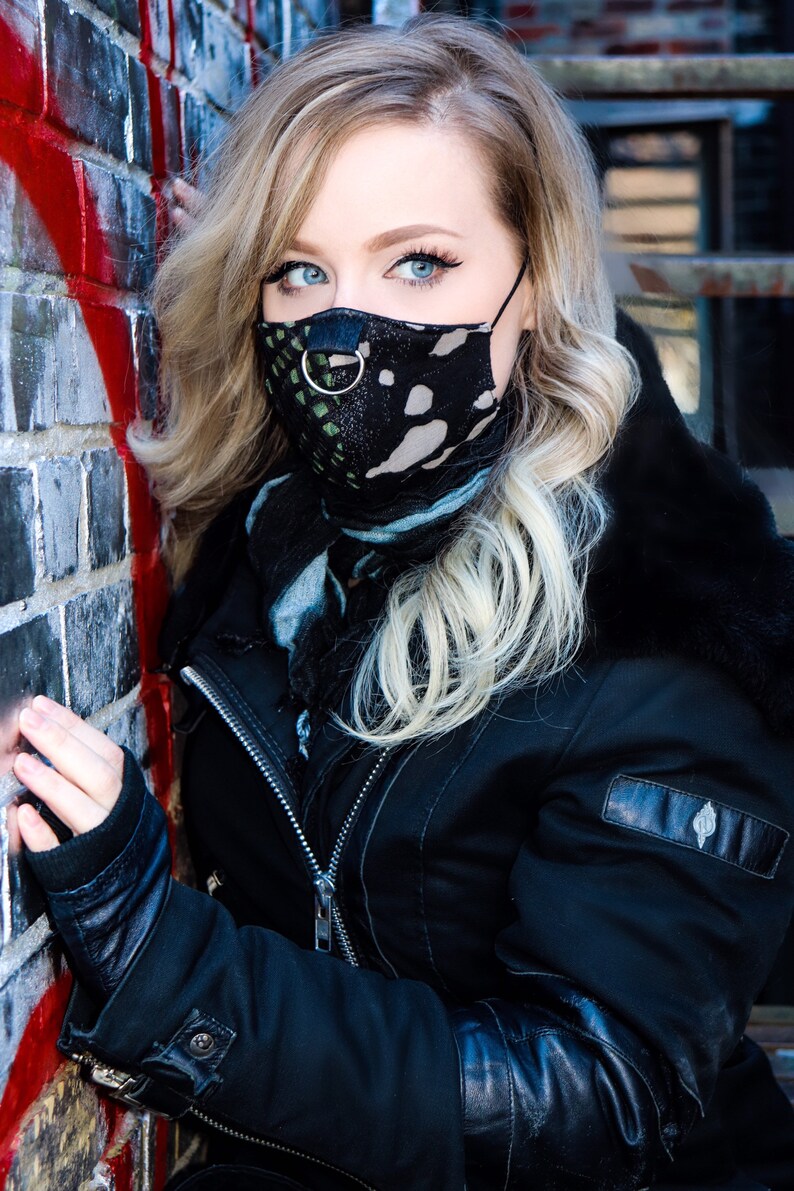 May include: A woman wearing a black leather jacket with a black and green patterned face mask. The mask has a silver ring attached to it.