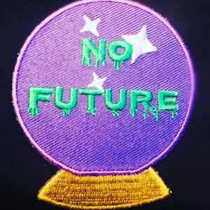 Può includere: Toppa in tessuto viola a forma di sfera di cristallo con base dorata. La toppa ha stelle bianche e testo verde che cola che dice "NO FUTURE".
