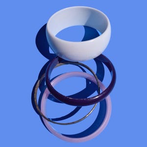 Puede incluir: Un conjunto de cuatro pulseras de plástico. La pulsera superior es azul claro, las dos siguientes son de color morado oscuro y la pulsera inferior es de color rosa claro.