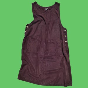Könnte beinhalten: Ein dunkelbraunes, ärmelloses Latzkleid mit rundem Ausschnitt und zwei Vordertaschen. Das Kleid hat Knopfverschlüsse an den Seiten und ist aus einem weichen, fließenden Stoff gefertigt. Das Kleid wird vor einem grünen Hintergrund gezeigt.