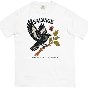 May include: A white short-sleeve t-shirt featuring a graphic of a black and white bird perched on an oak branch. Above the bird is the word SALVAGE in black, and below the branch is the text GATHER • MEND • REBUILD in black, with a red star.