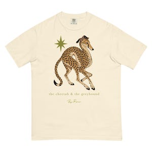May include: A cream-colored short-sleeve t-shirt featuring a graphic of a spotted cheetah in a running pose next to a gold star. Below the animal is the text "the cheetah & the greyhound" and a signature in gold script on the front center.