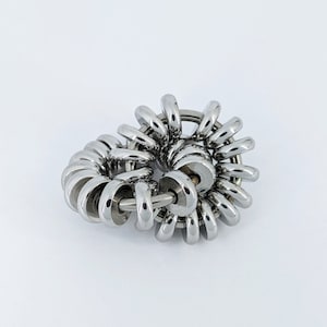 Op de afbeelding: Een zilverkleurig metalen fidget-speelgoed met meerdere in elkaar grijpende ringen. De ringen zijn in een spiraalvorm gerangschikt, waardoor een complex en ingewikkeld ontwerp ontstaat.