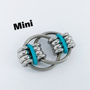 Pode incluir: Brinquedo anti-stress miniatura com dois anéis entrelaçados e bandas de borracha turquesa. Os anéis são de metal prateado.