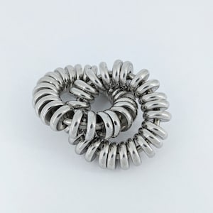 Puede incluir: Objeto met&aacute;lico plateado brillante compuesto por numerosos anillos circulares, dispuestos en un patr&oacute;n en espiral. Los anillos est&aacute;n estrechamente conectados, formando una estructura continua y enrollada. Fondo blanco.