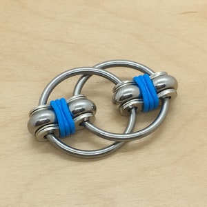 Op de afbeelding: Een zilveren metalen fidget-speelgoed met twee in elkaar grijpende ringen. De ringen zijn versierd met blauwe rubberen banden.